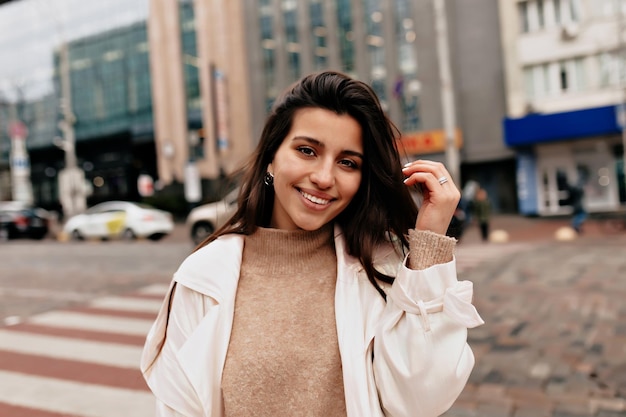 Retrato ao ar livre de encantadora mulher bonita com cabelo escuro e sorriso maravilhoso está andando na rua