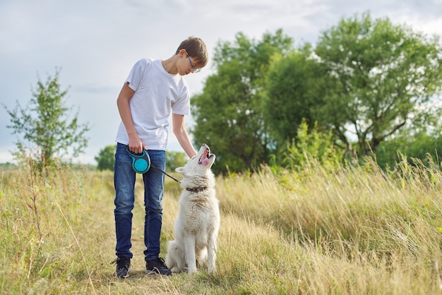 Retrato ao ar livre de menino com cachorro branco. Amizade de adolescente com animal de estimação, menino caminhando em um prado de verão, fundo de paisagens cênicas do sol, espaço de cópia