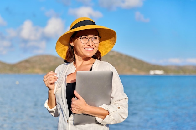 Retrato ao ar livre de mulher madura de chapéu na praia com um laptop nas mãos