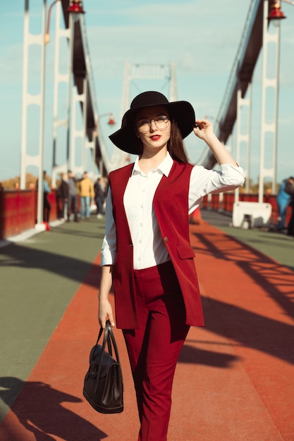 Retrato ao ar livre de uma jovem modelo elegante em traje vermelho e chapéu preto, posando na ponte em um dia ensolarado.