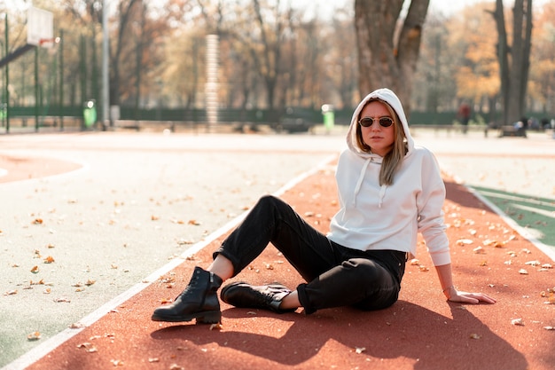 Retrato ao ar livre de uma jovem mulher bonita com long em óculos de sol e um suéter branco com capuz, sentado na pista de sportsground. cultura jovem passatempo de verão