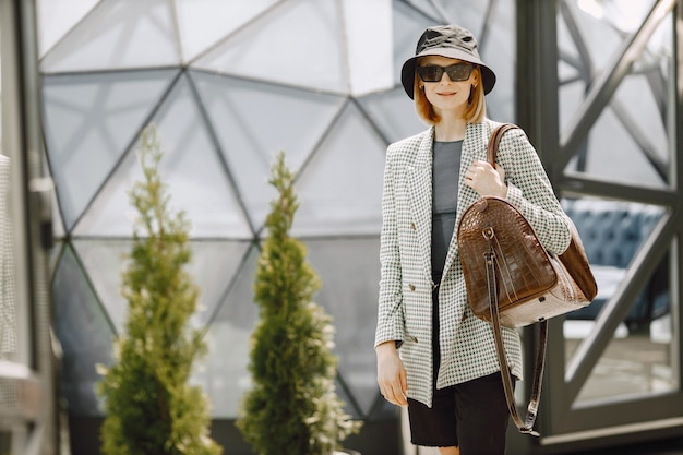 Retrato ao ar livre de uma jovem mulher bonita e elegante andando na rua, segurando uma grande bolsa de couro marrom. Rapariga loira com óculos escuros e chapéu preto