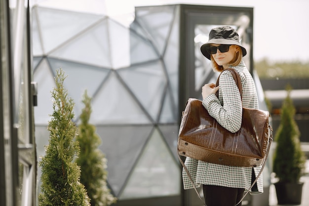 Retrato ao ar livre de uma jovem mulher bonita e elegante andando na rua, segurando uma grande bolsa de couro marrom. Rapariga loira com óculos escuros e chapéu preto