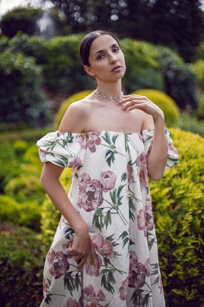 Retrato ao ar livre de uma linda mulher morena de luxo em um vestido com flores fica em um parque com árvores aparadas