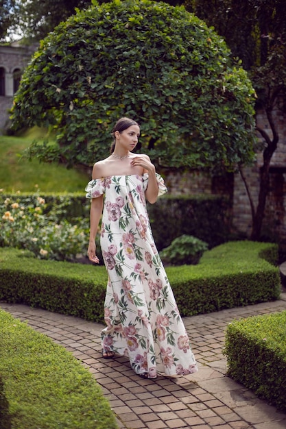 Retrato ao ar livre de uma linda mulher morena de luxo em um vestido com flores fica em um parque com árvores aparadas