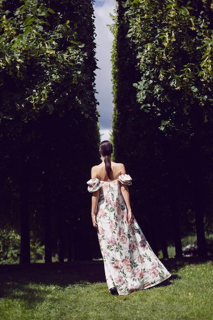 Retrato ao ar livre de uma linda mulher morena de luxo em um vestido com flores fica em um parque com árvores aparadas