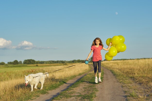 Retrato ao ar livre dinâmico de correr menina com cachorro branco e balões amarelos