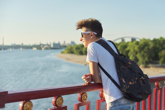 Retrato ao ar livre do jovem adolescente sorridente com um corte de cabelo na moda em pé na ponte sobre o rio em um dia ensolarado de verão, copie o espaço