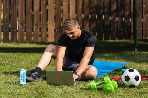 Retrato ao ar livre do jovem em um uniforme esportivo está descansando, espreguiçando-se no gramado, assistindo a um filme e estudando em um laptop, uma rede social no jardim