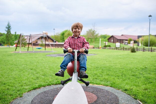 Retrato ao ar livre do menino risonho pré-escolar fofo balançando em um balanço no parque infantil