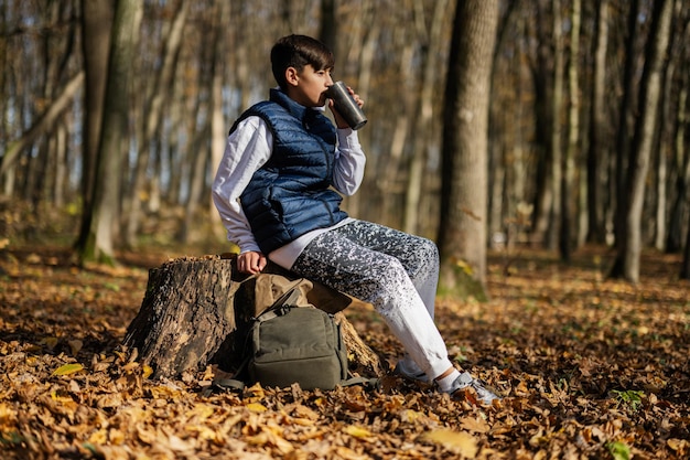 Retrato ao ar livre do outono de um adolescente sentado no toco e bebendo chá da xícara térmica na floresta