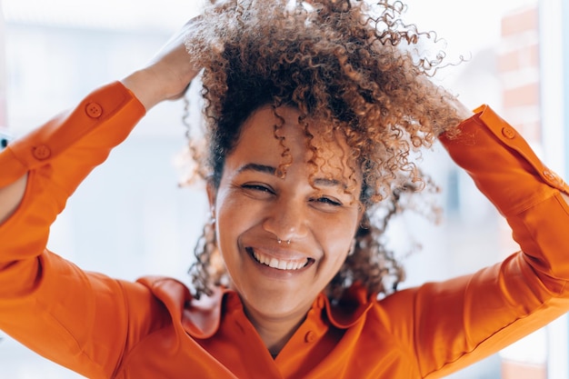 Retrato aproximado de cabelo encaracolado sorrindo jovem brasileira de camisa laranja com piercing no nariz com as mãos no cabelo