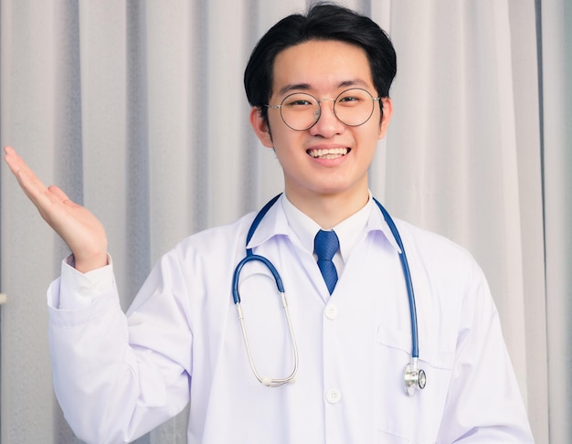 Retrato aproximado de feliz jovem médico asiático, homem bonito, sorrindo em uniforme e alça de pescoço de estetoscópio, mostrando as mãos e olhando para a câmera com espaço de cópia, conceito de medicina de saúde