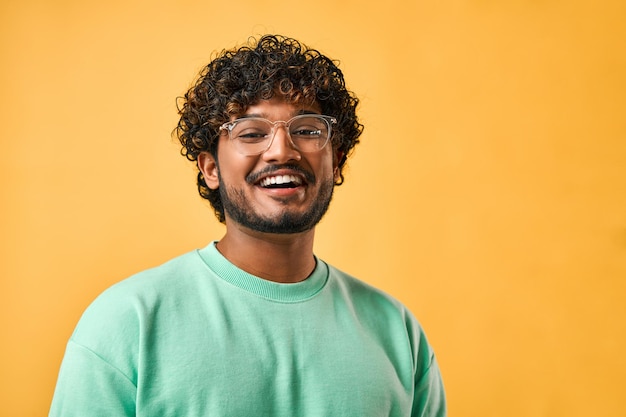 Retrato aproximado de um belo homem indiano de cabelos encaracolados em uma camiseta turquesa e óculos rindo e olhando para a câmera