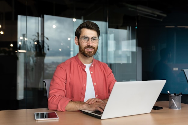 Retrato aproximado de um empresário maduro dentro de um escritório com barba e camisa vermelha sorrindo e