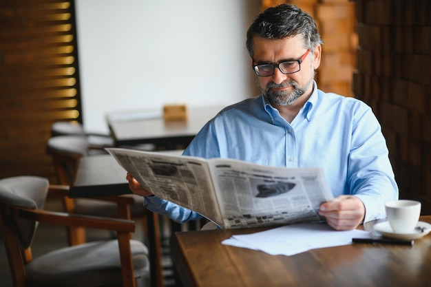 Retrato aproximado de um homem bonito sênior sério lendo jornal tomando café e sentado à mesa