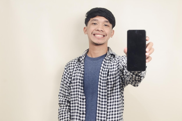 Retrato aproximado de um jovem modelo asiático feliz vestindo camisa xadrez apontando para a tela do telefone em fundo isolado