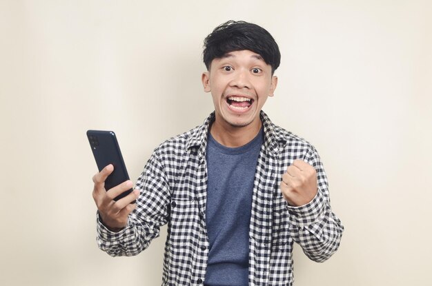 Retrato aproximado de um jovem modelo asiático feliz vestindo camisa xadrez com expressão vencedora enquanto segura o telefone