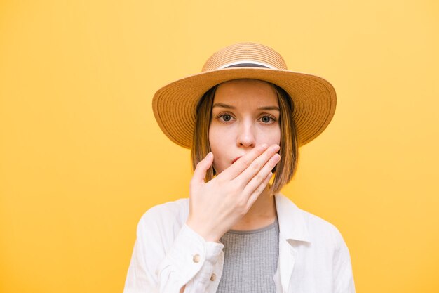 Retrato aproximado de uma garota chocada de chapéu em fundo amarelo olhando para a câmera com a boca coberta