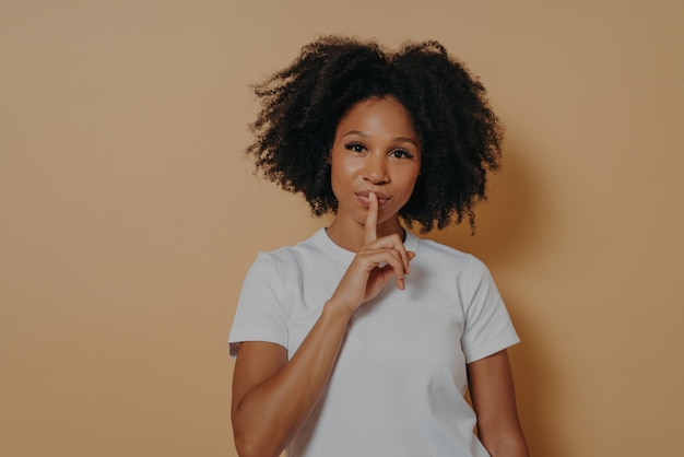 Retrato aproximado de uma jovem afro-americana de cabelos ondulados em camiseta branca mostrando sinal de shh