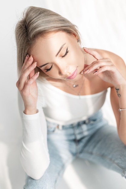 Retrato aproximado de uma linda jovem loira com olhos verdes e um rosto bonito em uma camiseta branca em um fundo branco com luz do sol