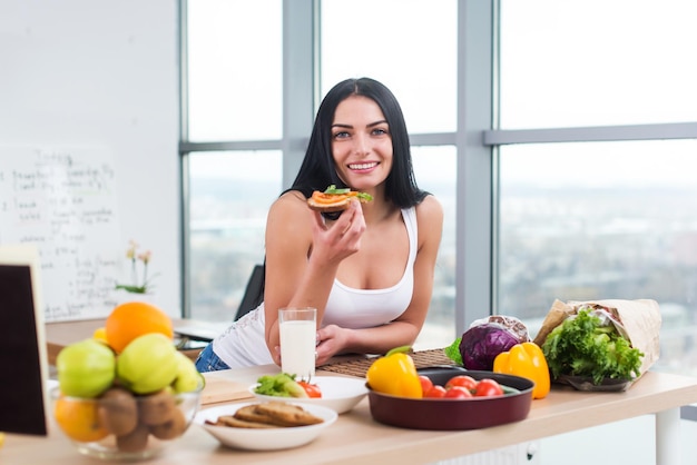 Retrato aproximado de uma mulher sorridente comendo sanduíche vegetariano dietético com legumes no café da manhã olhando para a câmera