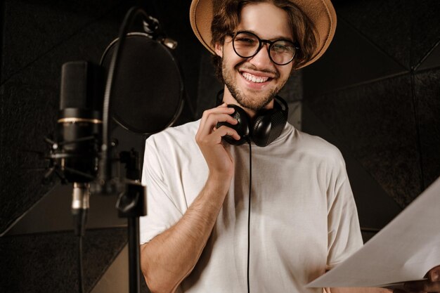 Retrato de un apuesto cantante masculino barbudo mirando felizmente a la cámara con el texto de una nueva canción en un moderno estudio de grabación de sonido