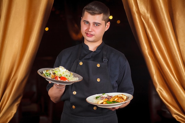 Retrato de un apuesto chef sonriente en uniforme de pie con un delicioso plato en la cocina. Chef masculino sosteniendo plato con plato cocinado.