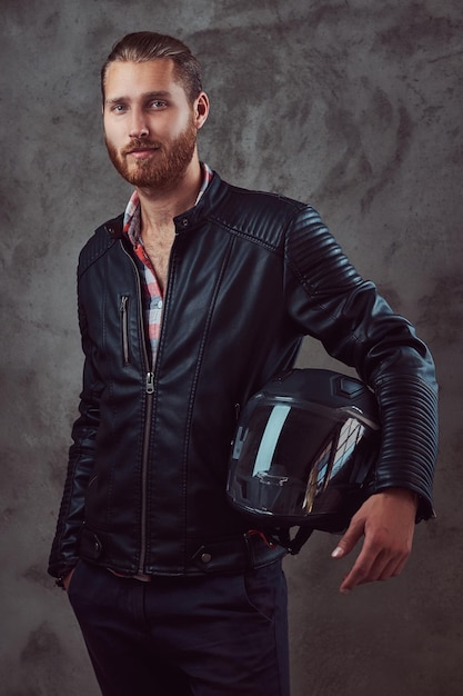 Retrato de un apuesto y elegante ciclista pelirrojo con una chaqueta de cuero negro, sostiene casco de motocicleta, posando en un estudio. Aislado en un fondo oscuro.