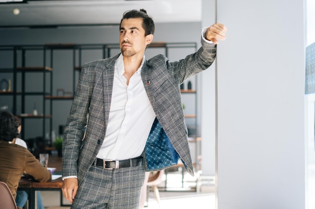 Retrato de un apuesto y elegante hombre de negocios con traje de moda parado junto a la ventana en una oficina moderna mirando hacia otro lado