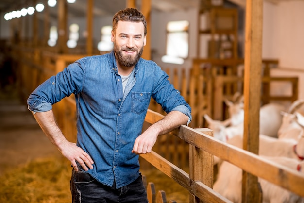 Retrato de un apuesto granjero de pie en el establo con cabras. Producción y cultivo de leche natural