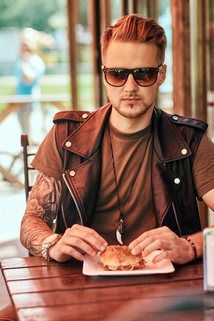 Retrato de un apuesto hipster con estilo se sienta en una mesa, pidió una hamburguesa y un refresco, cena en un café al borde de la carretera