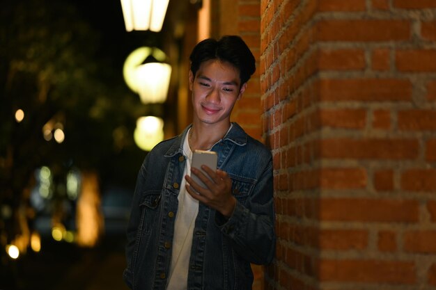 Retrato de un apuesto hombre asiático escribiendo un mensaje de texto en un teléfono inteligente mientras está de pie en las calles de la ciudad nocturna