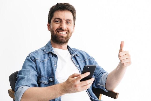 Retrato de un apuesto hombre barbudo con ropa casual sentado en una silla aislada sobre una pared blanca, sosteniendo un teléfono móvil