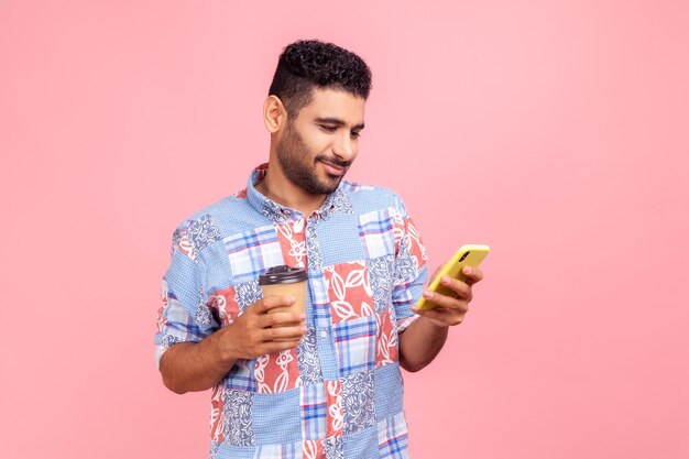 Retrato de un apuesto hombre barbudo satisfecho con un pantalones azul de estilo casual de pie con café y charlando por teléfono expresando emociones positivas Camisa de estudio interior aislada en un fondo rosa