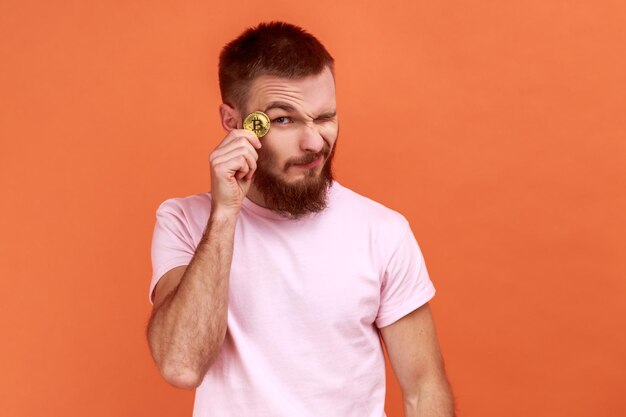 Retrato de un apuesto hombre barbudo sonriente sosteniendo bitcoin cerca del ojo, satisfecho con una moneda btc dorada, dinero electrónico, usando una camiseta rosa. Disparo de estudio interior aislado sobre fondo naranja.