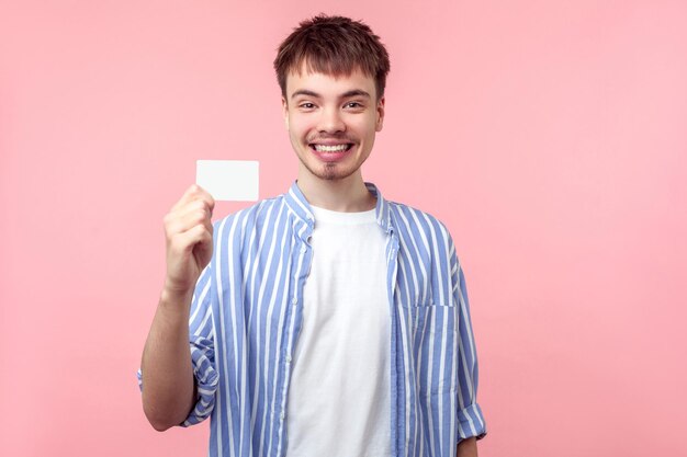 Retrato de un apuesto hombre de cabello castaño con barba pequeña y bigote en un pantalones informal que sostiene una tarjeta bancaria de plástico blanca y sonríe a la cámara. tiro de estudio interior aislado sobre fondo de color rosa