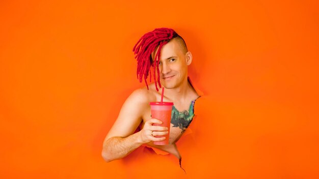 Retrato de un apuesto hombre sin camisa con rastas rojas y un tatuaje de un pájaro en el pecho bebiendo refrescos o jugo a través de una pajita mirando la cámara a través de un agujero en la pared naranja