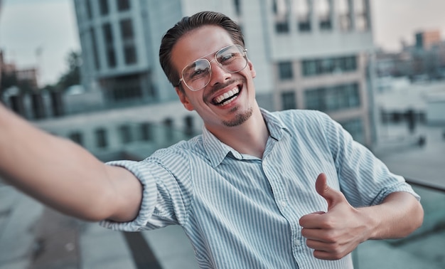 Retrato de un apuesto hombre feliz haciendo el signo de ok y tomando un selfie al aire libre con su teléfono inteligente.