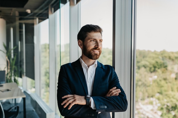 Retrato de un apuesto hombre de negocios caucásico de pie con los brazos cruzados cerca de la ventana con un descanso en el espacio libre de la oficina