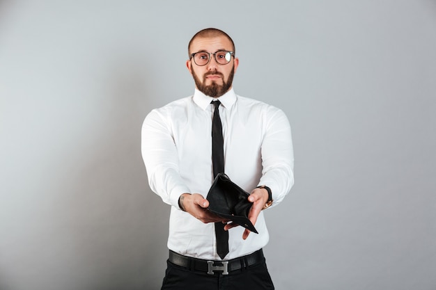 Retrato de apuesto hombre de negocios molesto en gafas y traje mostrando su billetera vacía, aislado sobre la pared gris