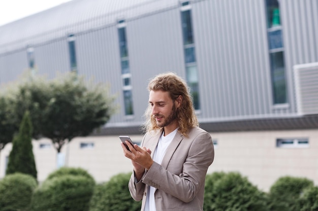 Retrato de un apuesto hombre de negocios rizado con ropa informal que sostiene un teléfono inteligente y una gestión exitosa sonriente