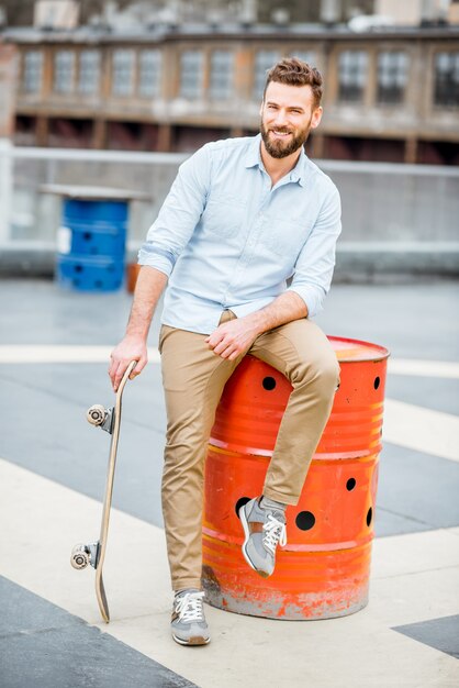 Retrato de un apuesto hombre de negocios sentado con patineta en el patio de la azotea en el edificio industrial