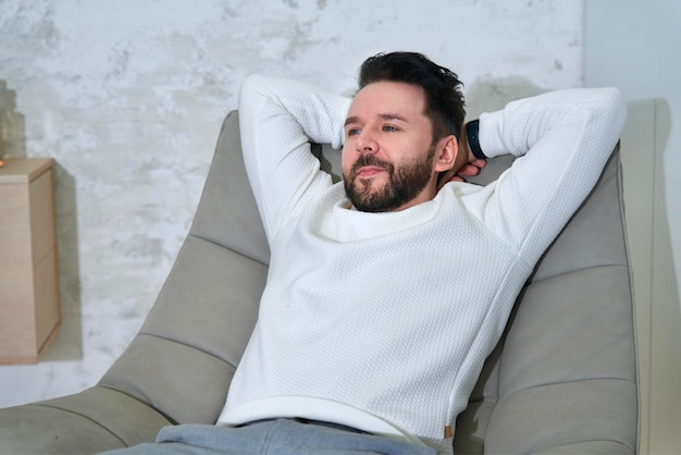 Retrato de un apuesto hombre relajado y exitoso, el joven está sentado en un cómodo sillón en su lujoso piso, apartamento o casa, relajándose, descansando, descansando, soñando, pensando, disfrutando de la vida