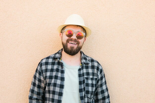 Retrato apuesto hombre vestido con sombrero de verano y gafas de sol y camisa a cuadros sonriendo feliz cerca de la pared trav