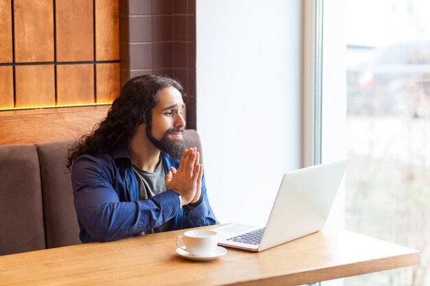 Retrato de un apuesto joven adulto independiente preocupado con estilo informal sentado en un café y hablando con su amigo en una computadora portátil cogido de la mano como rezar y disculparse Concepto de estilo de vida interior