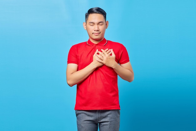 Retrato de un apuesto joven asiático con camisa roja presionando las manos contra el pecho con los ojos cerrados y un gesto agradecido en la cara. concepto de salud.