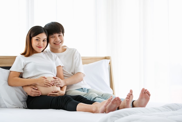 Retrato de un apuesto joven asiático y una mujer vestida con un camisón blanco sentados juntos en una cama. Están sonriendo felices y tocando el vientre de una madre embarazada.