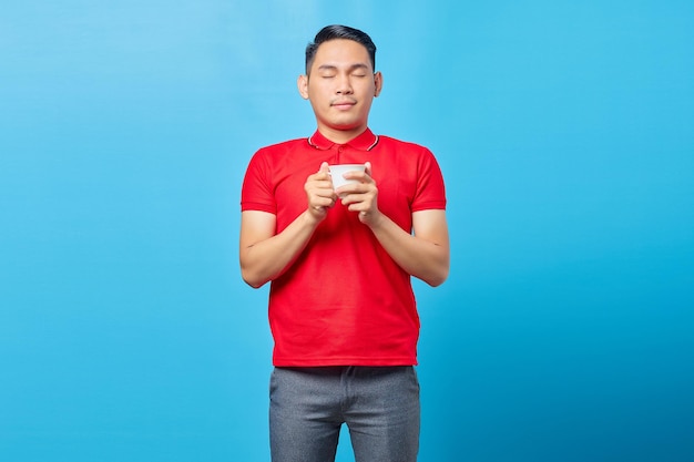 Retrato de un apuesto joven asiático sosteniendo una taza de café y cerrando los ojos mientras inhalaba un delicioso aroma de café aislado de fondo azul