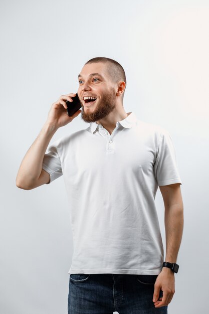 Retrato de un apuesto joven en una camiseta blanca hablando por teléfono inteligente sobre fondo blanco.